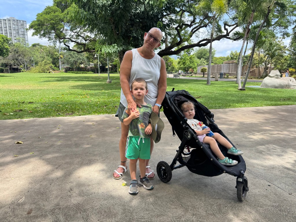 A woman stands smiling with two young children in front of a green lawn