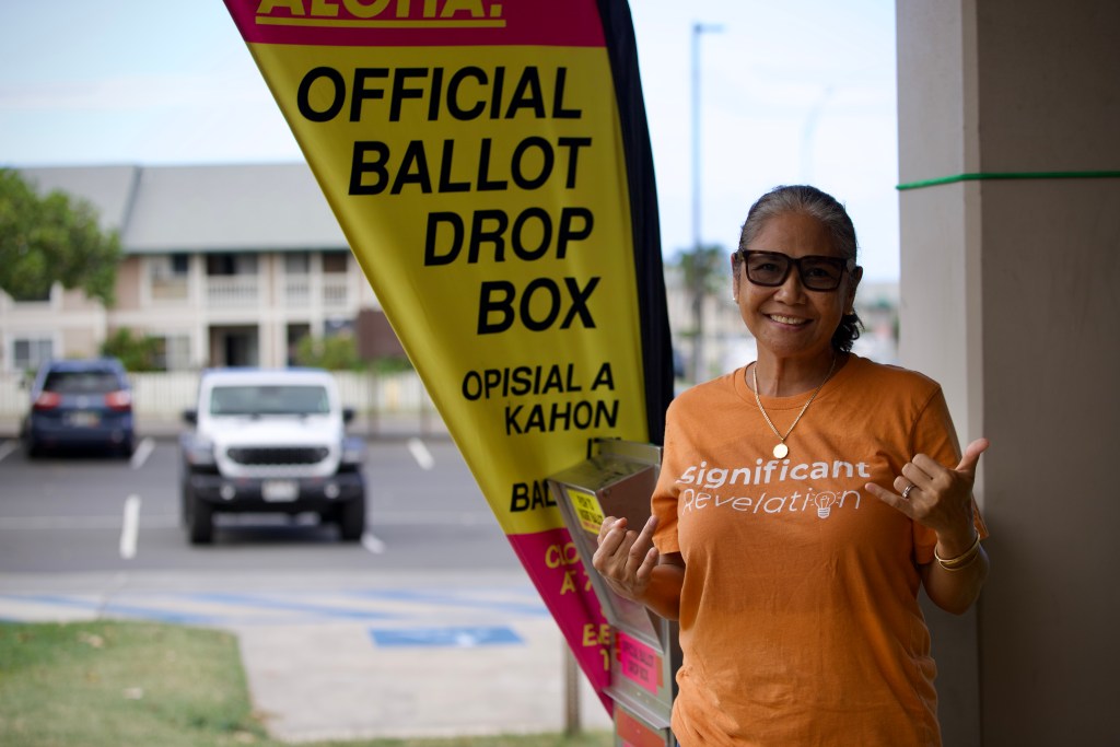 Angelina Abapo, wearing an orange t-shirt and her hair in a ponytail, smiles and stands in front of the official voting dropbox in front of the Velma McWayne Santos Community Center.