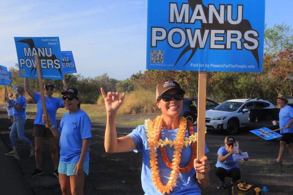 A woman wearing leis, sunglasses, and a hat holds an election sigh on the side of the road, surrounded by supporters in matching t-shirts.