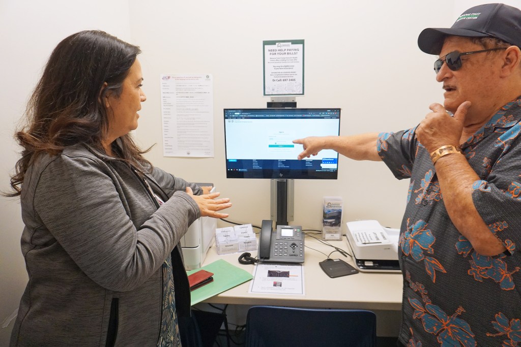 Waianae Coast Comprehensive Health Center CEO Rich Bettini (right) and Vice President Leinaala Kanana demonstrate how to use pods throughout the campus that connect patients via phone to an employee that will help them submit needed information and applications to the state’s Medicaid program. (Ashley Mizuo/KFF Health News)