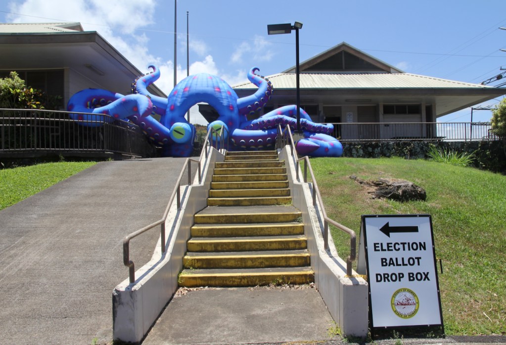 An giant air-filled squid sits beside a sign notifying the public of the location of a ballot drop box.