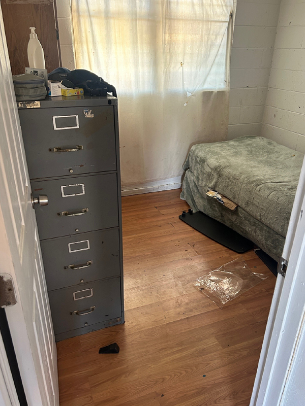A small room with a bed,a gray filing cabinet and white curtains with holes is viewed from the doorway.