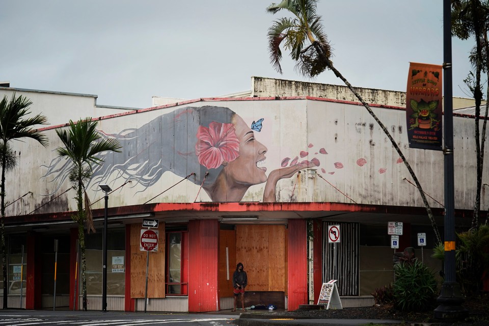 A homeless man stands under a mural of a wahine with wind-blown hair and flower petals blowing as Hawaiʻi Island prepares for Hurricane Nolo Friday, Sept. 25, 2026, in downtown Hilo. (Kevin Fujii/Civil Beat/2026)
