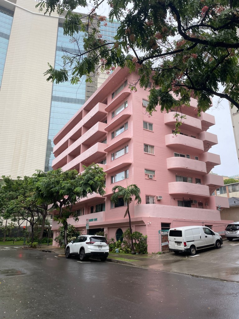 Pink stucco building with white cars in the foreground photographed from the street shown on an angle.