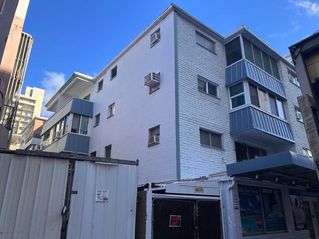 A white and grey multistory building seen from the street showing the corner of the building and the windows to several units.