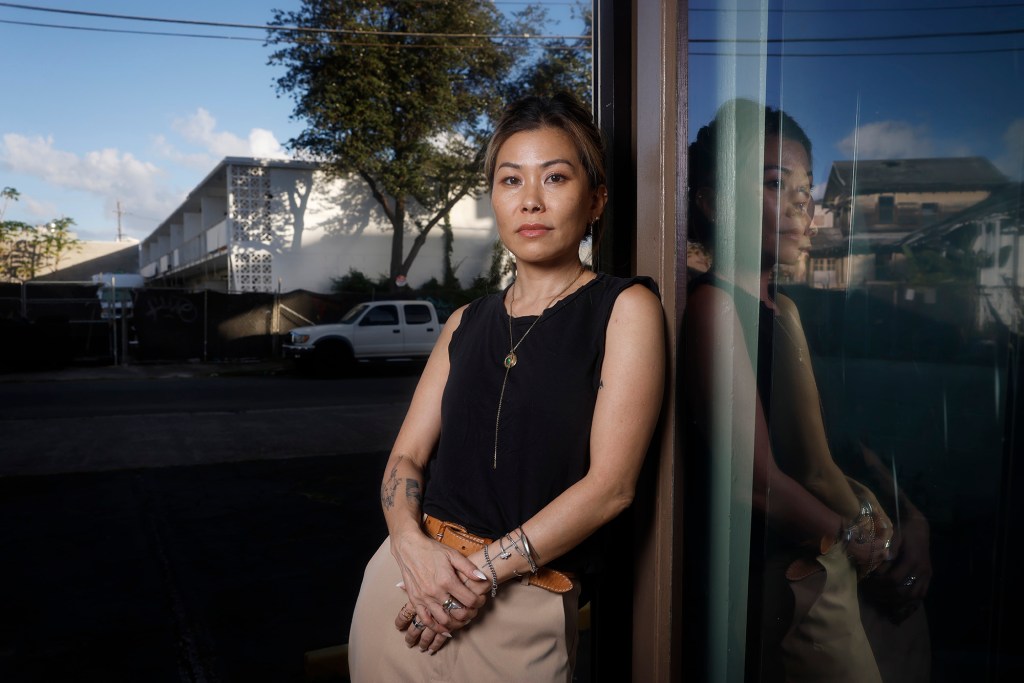 Emiko Miyazawa stands in front of 929 Pumehana Street Wednesday, Oct. 21, 2026, in Honolulu. The property contains multiple buildings in various stages of ruin, two of which are reflected in the glass at right. (Kevin Fujii/Civil Beat/2026)