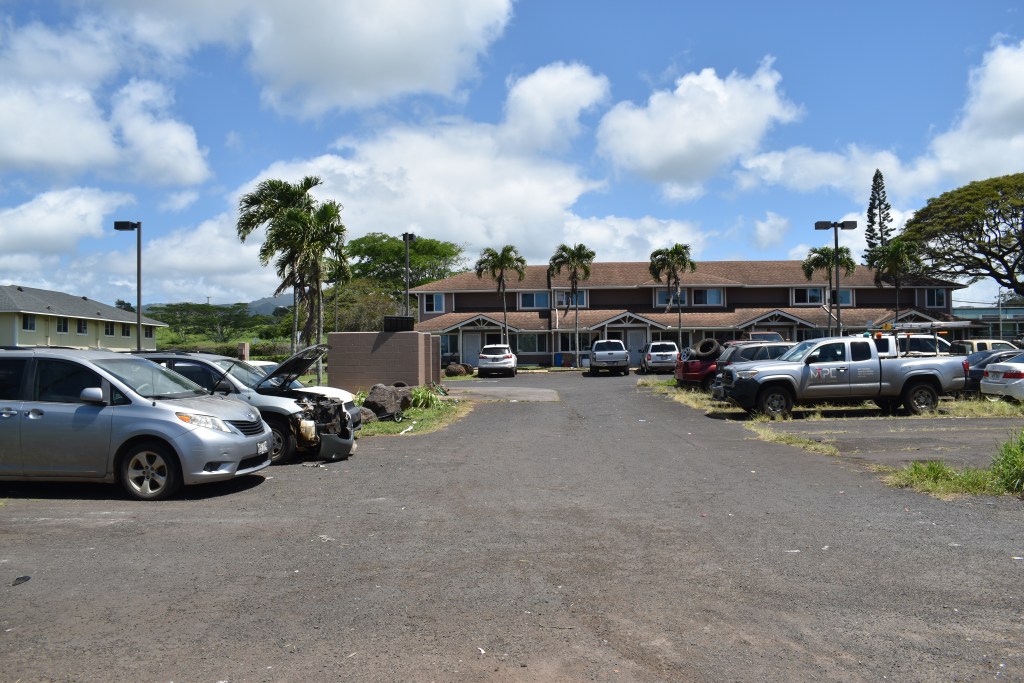 A half full parking lot at the Kawailehua Public Housing Complex in Kōloa, Kaua‘i with an abandoned and partially stripped car and two-story tan building in the background.